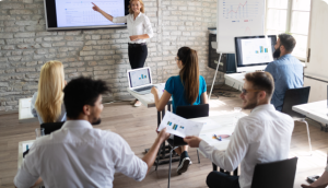 "A staffing agency executive providing guidance to recruiters and account managers on industry best practices. The executive is standing in front of a group of attentive professionals, engaging them in a discussion. The room is well-lit with a professional atmosphere, and everyone is focused on the executive's presentation. The executive is gesturing confidently, emphasizing key points while holding a visual aid that illustrates staffing strategies. The atmosphere reflects a collaborative and productive environment for learning and improving skills within the staffing industry."