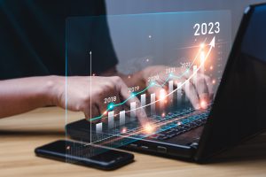 A pair of hands positioned on a laptop keyboard, typing while a holographic chart hovers above the keyboard. The hands are delicately tying and pressing the keys, while the holographic chart displays a visually captivating representation of data and graphs, emitting a futuristic and dynamic glow.